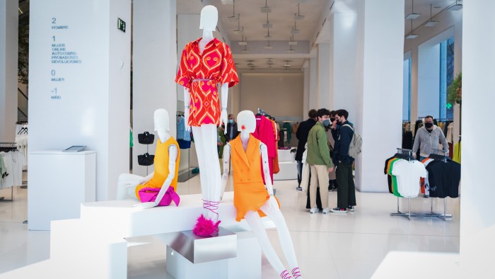 a view of the interior of a fashion store. There are dressed mannequins in the foreground. At the back are people standing in line